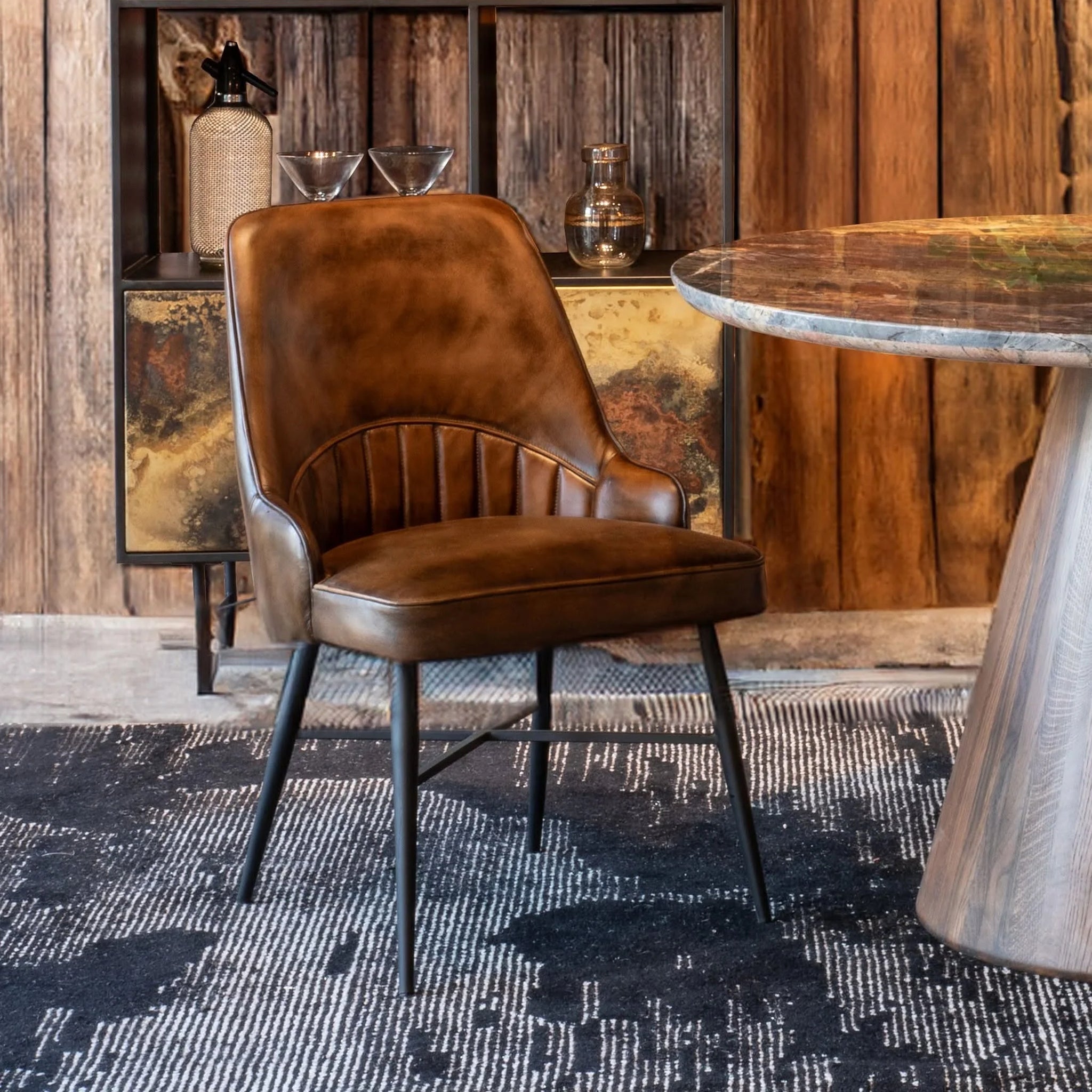 A brown leather dining chair with black iron legs, positioned in a rustic room setting with a wooden wall, a patterned rug, and a stone-topped table.