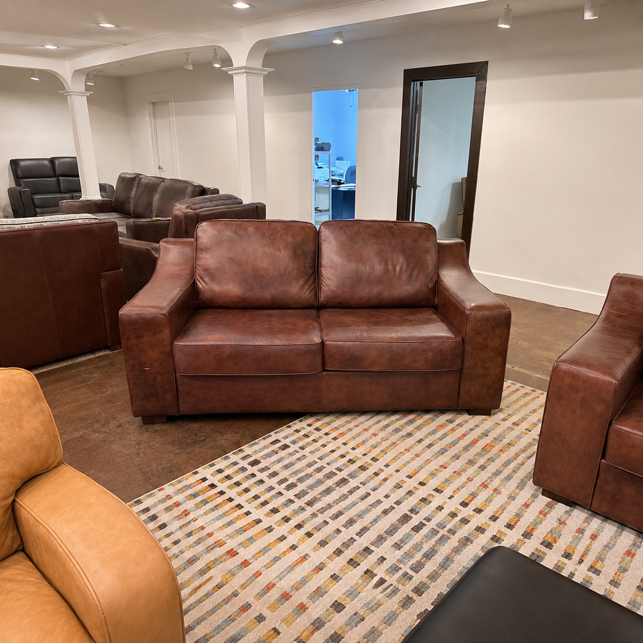 A brown leather loveseat sofa in a showroom setting, surrounded by other leather furniture pieces and a patterned area rug.