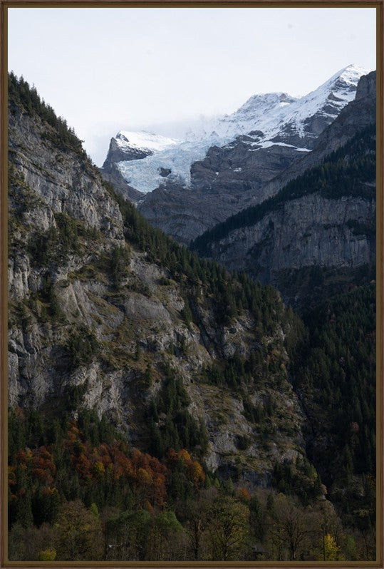 A framed landscape photograph of a dramatic mountain valley, featuring steep rocky cliffs, evergreen forests, and snow-capped peaks under a pale sky.