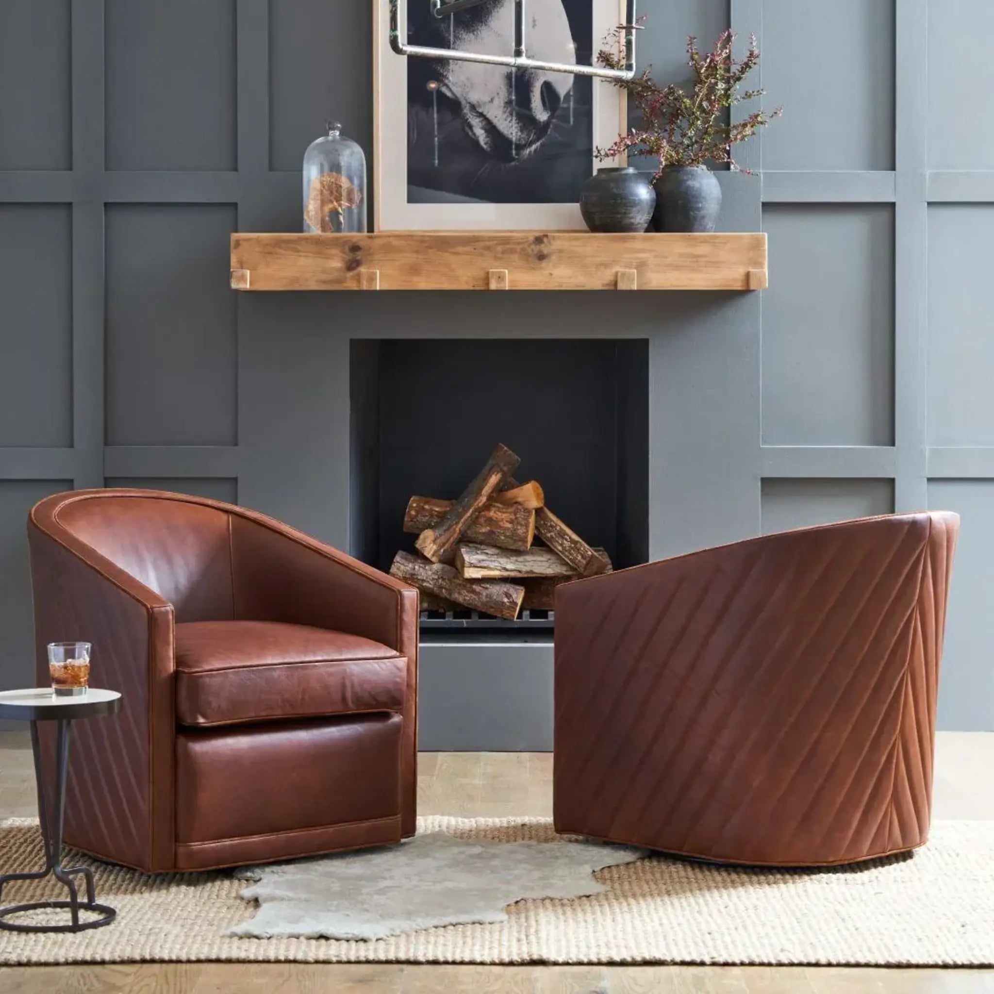 Two brown leather swivel armchairs in a styled living room setting with a fireplace, gray paneled wall, and a small side table.