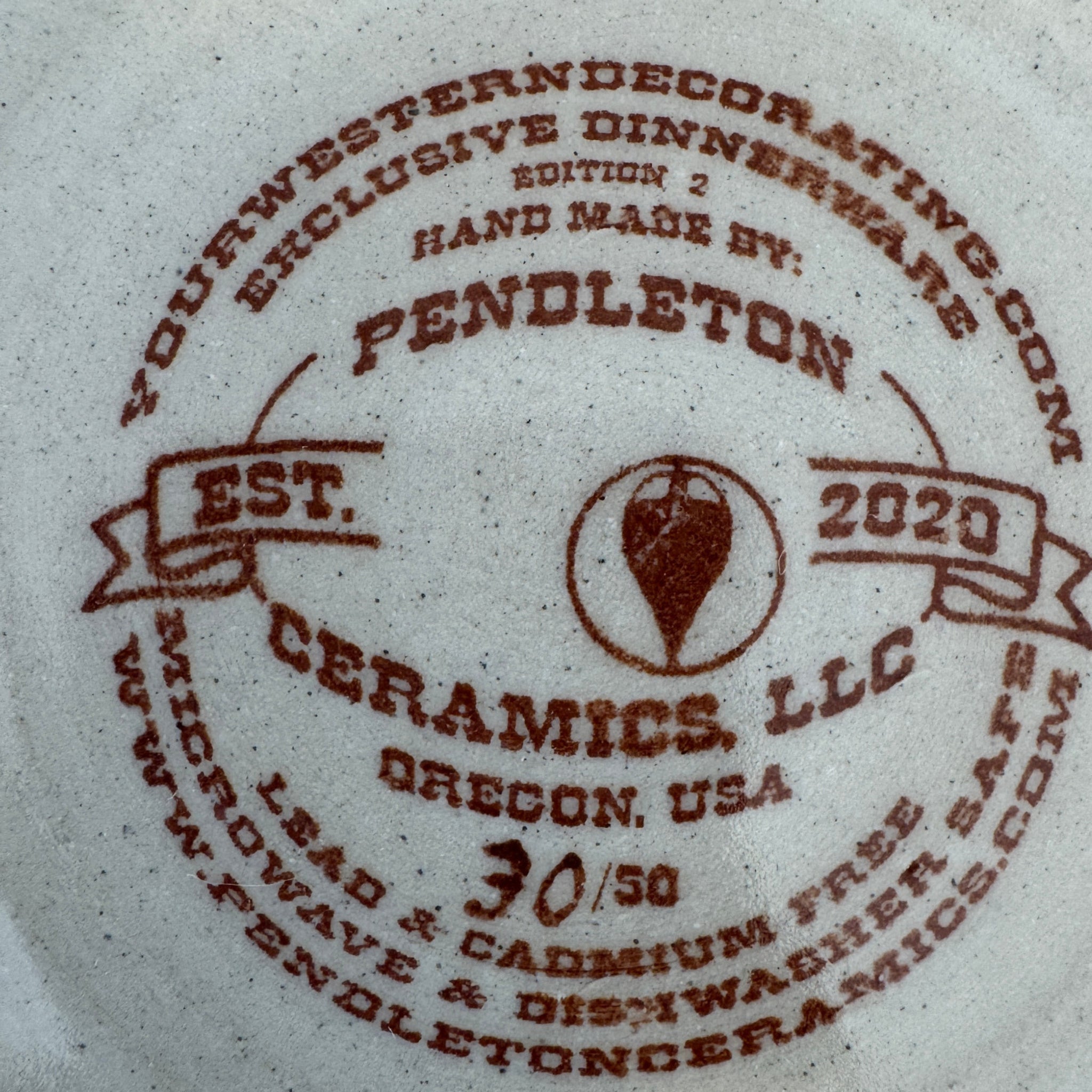 Close-up of a white ceramic dinnerware piece printed with a brown Pendleton Ceramics LLC logo and text, including 'Exclusive Dinnerware' and 'Hand Made By: Pendleton Ceramics, LLC. Exclusive Dinnerware for Your Western Decor