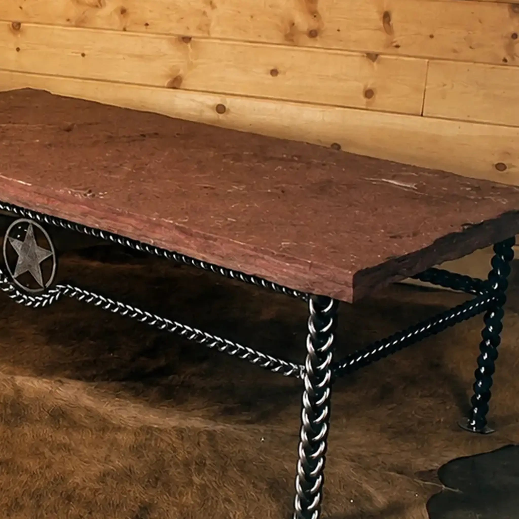 A rustic western-style table with a thick reddish-brown stone top and a black metal frame featuring braided iron legs and a star emblem on the side, set against a wood-paneled wall and cowhide rug.