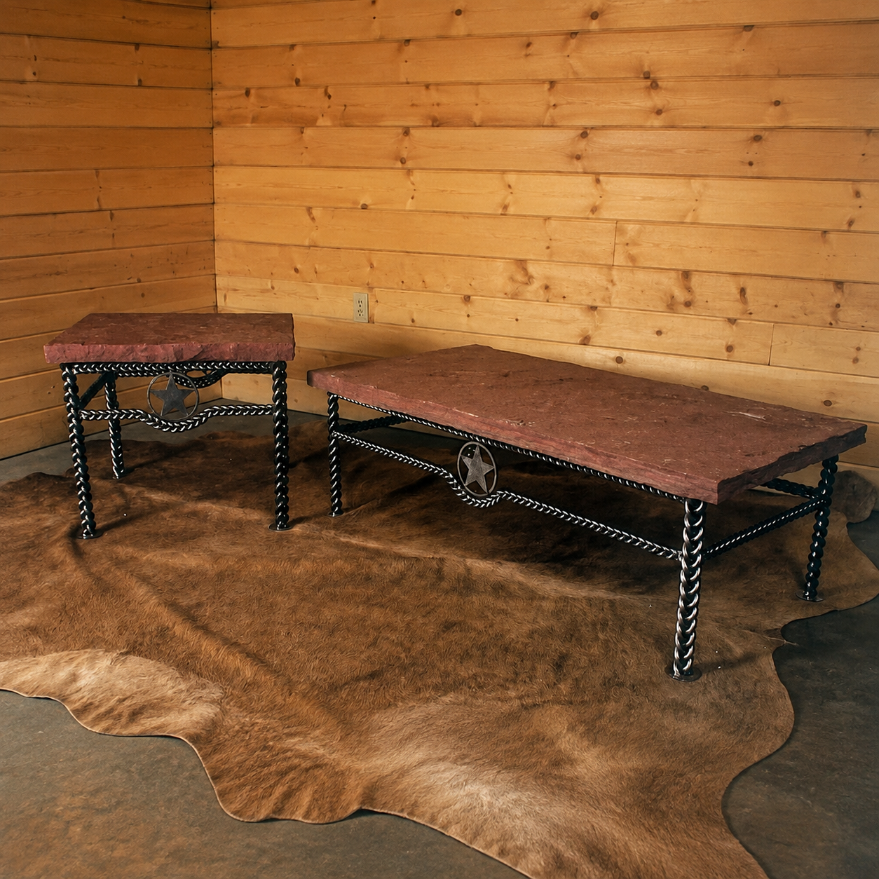 Two rustic western-style tables with reddish-brown stone tops and black metal legs with signature braided iron and center western star, displayed on a brown cowhide rug against a wood-paneled wall.