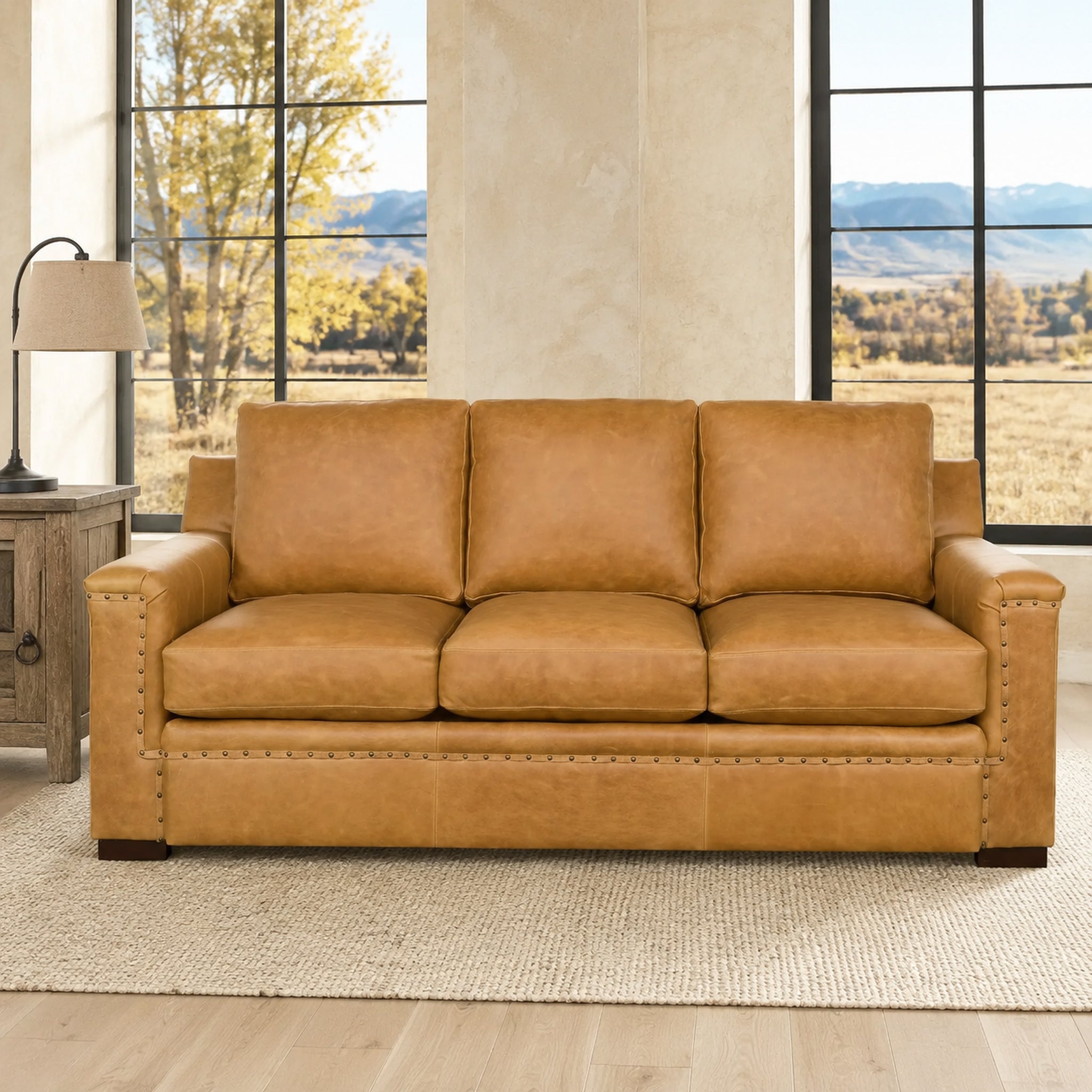 A three-seat leather sofa in a warm tan/caramel color, styled in a bright living room with large windows, a side table, and a lamp.