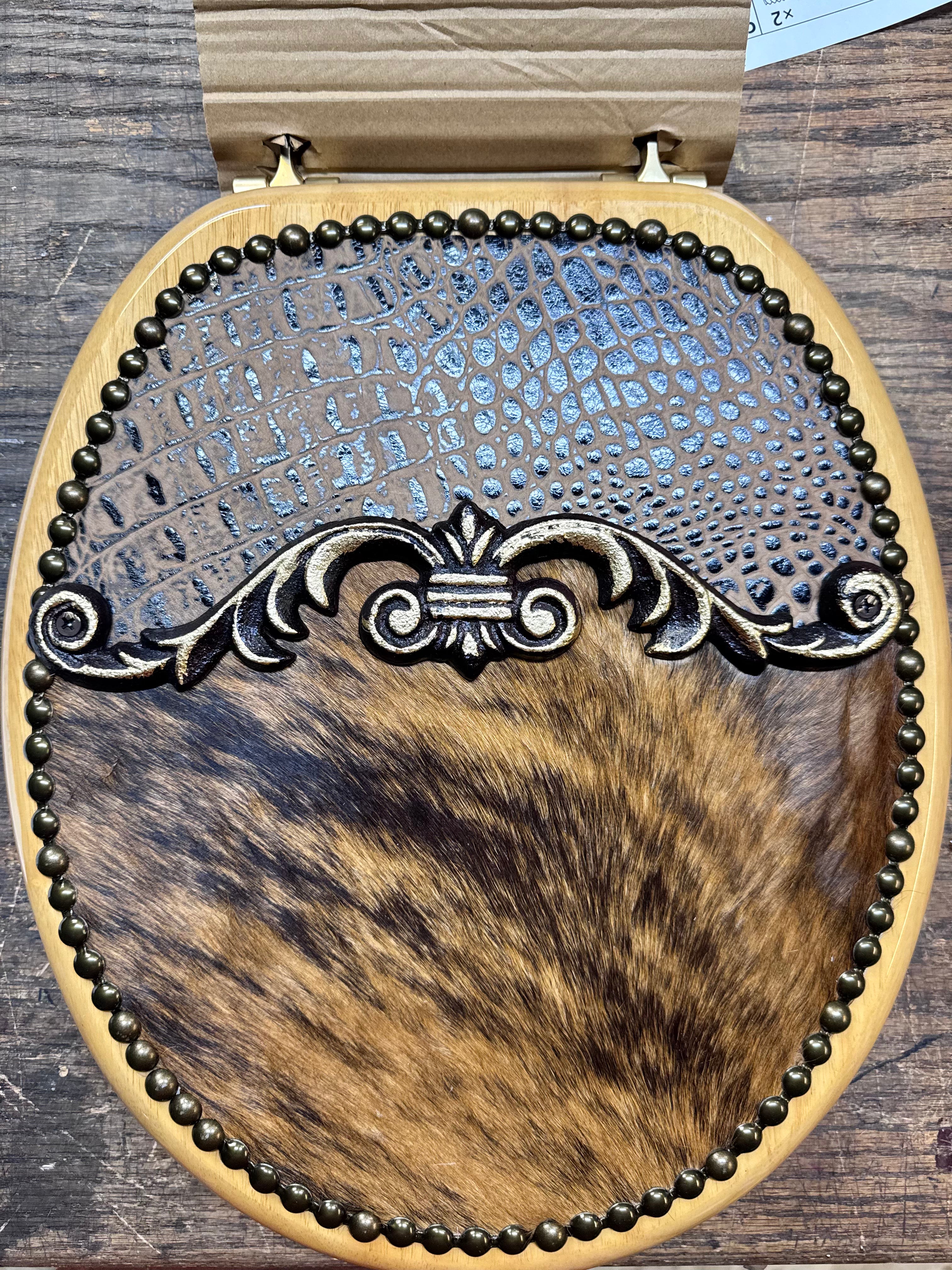 Western round toilet seat upholstered with brown croc embossed leather, brindle cowhide, with nail trim and decorative center design.