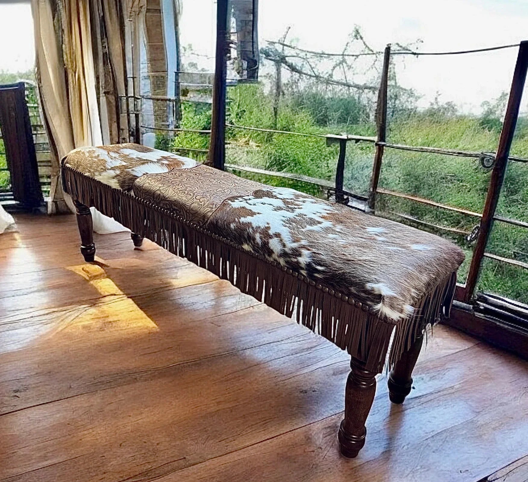 American made Wooden bench with cowhide cover and fringe on a wooden floor, large window in the background.