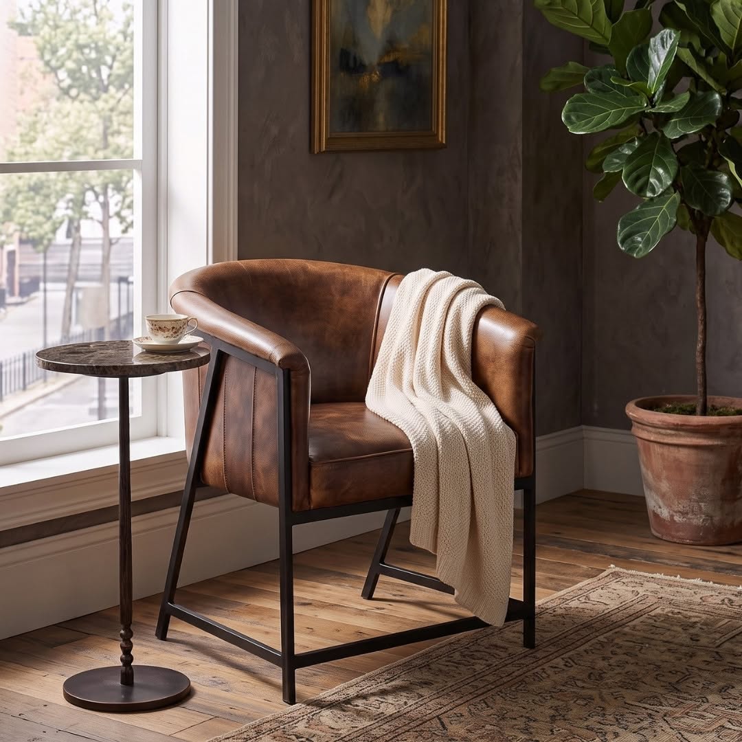 A brown leather barrel-style accent chair with a black metal frame, styled in a room with a cream knit throw blanket draped over it, a small round side table, a potted fiddle-leaf fig plant, and a patterned rug.