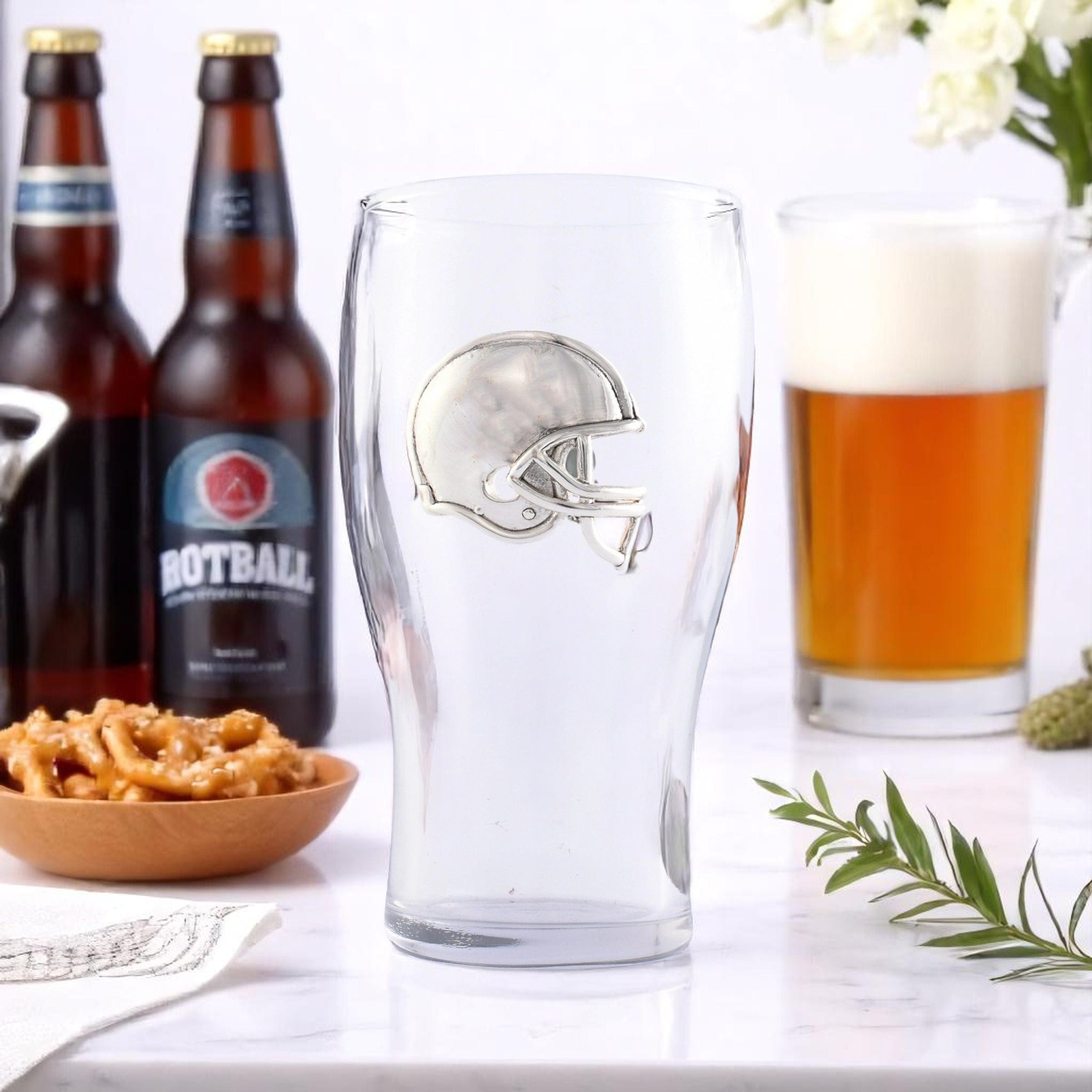 Clear glass with football helmet design, bottles of beer, pretzels, and a glass of beer on a white surface.