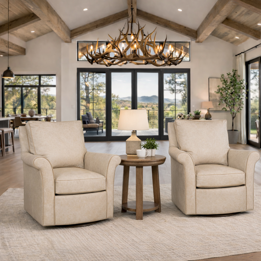 Two beige leather swivel chairs with rolled arms sit side by side in a bright luxury ranch living room, styled with a round wood side table and lamp between them, beneath exposed wood beam ceilings and an antler chandelier overlooking mountain views through black-framed glass doors.