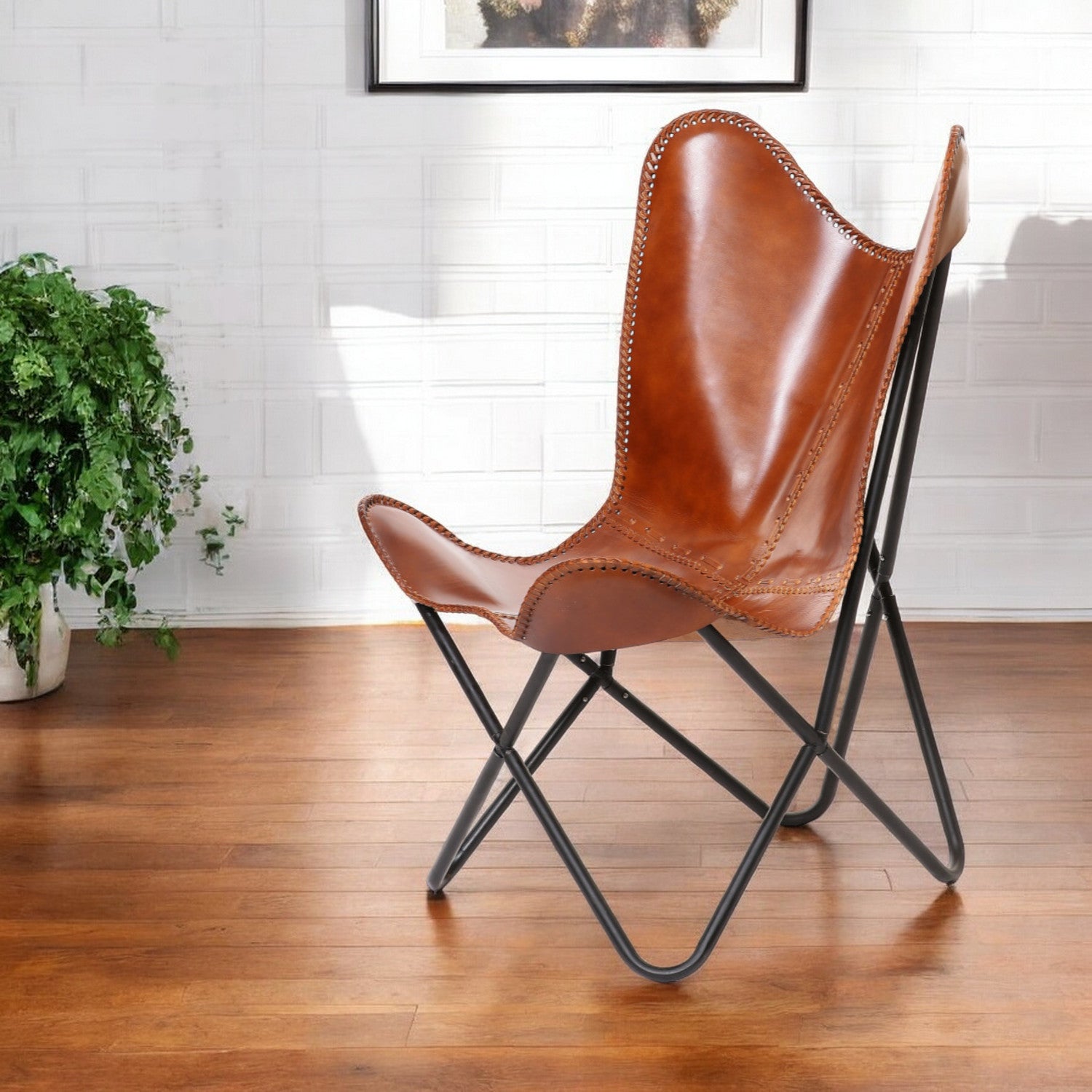 Brown leather butterfly chair on a wooden floor with a plant and framed picture in the background - Your Western Decor