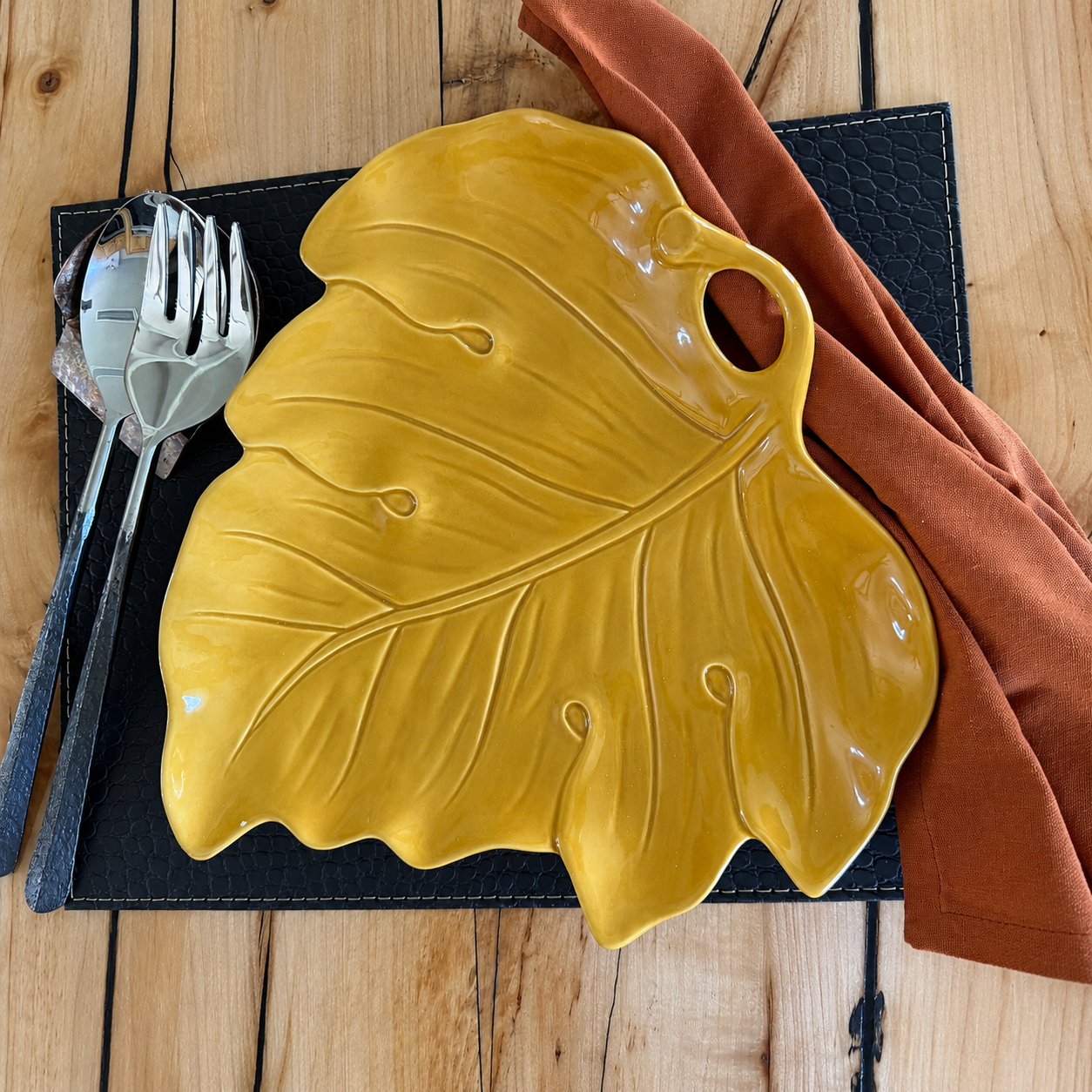 A glossy yellow ceramic leaf-shaped serving platter is styled on a black textured placemat with silver cutlery and a rust-orange napkin on a light wood table.