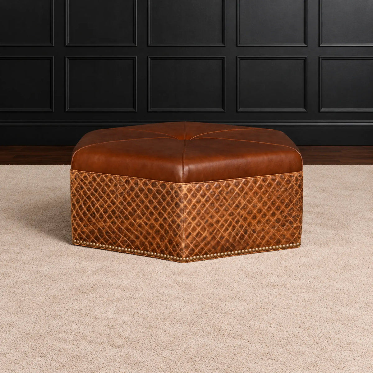 A brown leather ottoman with a diamond-patterned lower skirt and brass nailhead trim, set on a beige carpet in front of a black paneled wall.