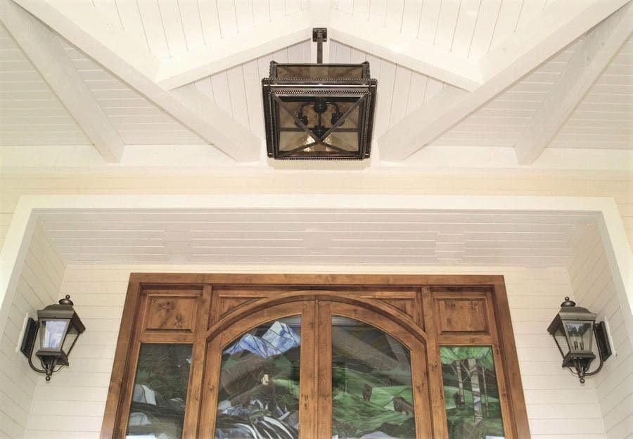 A square black metal outdoor pendant light hangs from the center of a white shiplap ceiling above a wooden double door with glass panels, flanked by two matching wall lanterns.