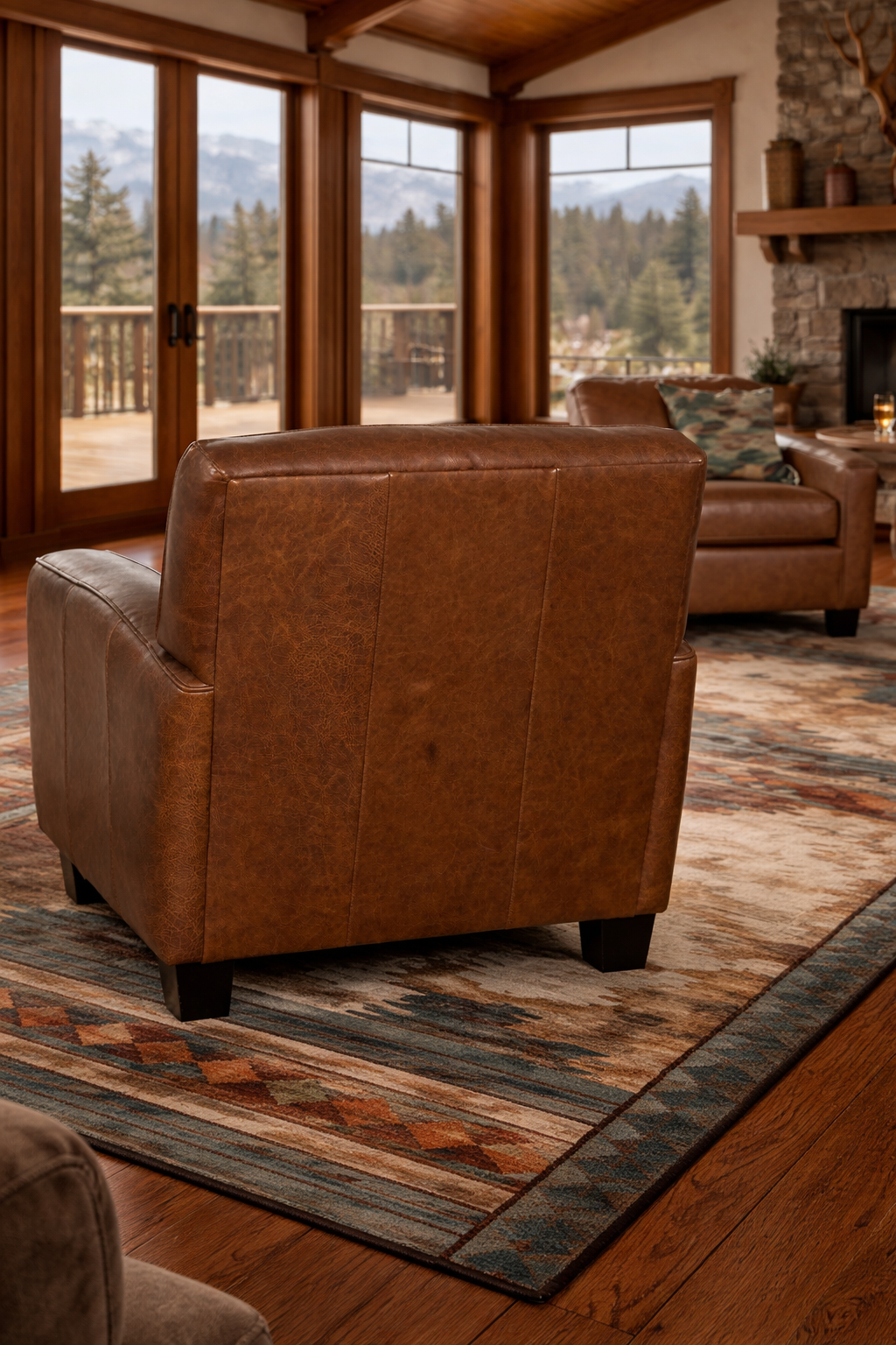 Back view of saddle brown full-grain leather club chair showcasing tailored panel stitching and rich artisan leather grain, styled in a luxury mountain lodge living room with Southwestern area rug, wood beam ceilings, and large windows overlooking mountain scenery.