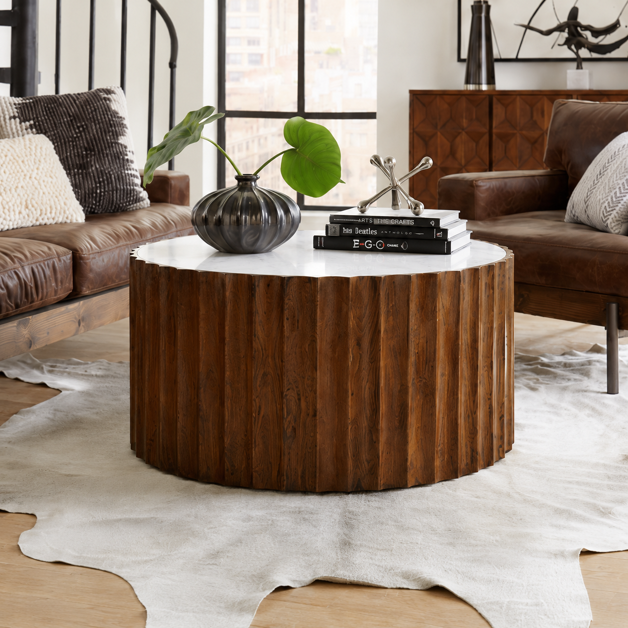 A round coffee table with a fluted dark wood base and a white top, styled in a modern living room with leather sofas, a cowhide rug, and decorative objects on top.