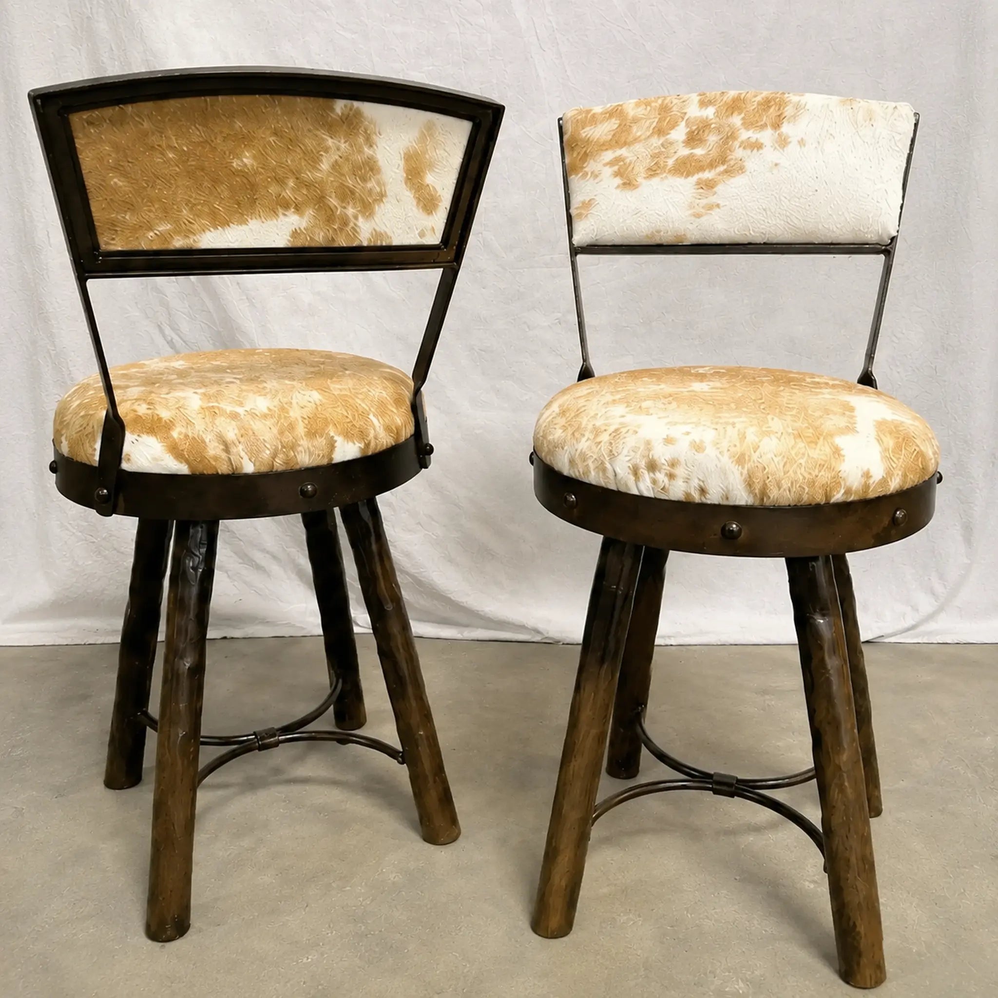 Two rustic bar stools with dark brown metal legs, metal frames, and cowhide upholstered seats and backrests, shown against a white draped backdrop.