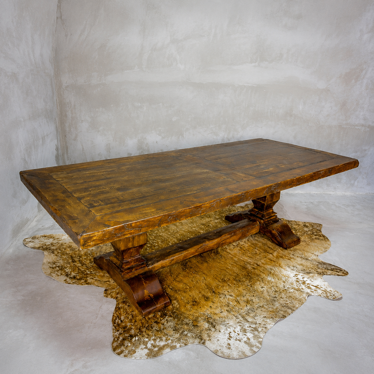 A rustic dark brown wooden table with a thick rectangular top and carved trestle-style legs, styled on a cowhide rug against a textured gray studio backdrop.