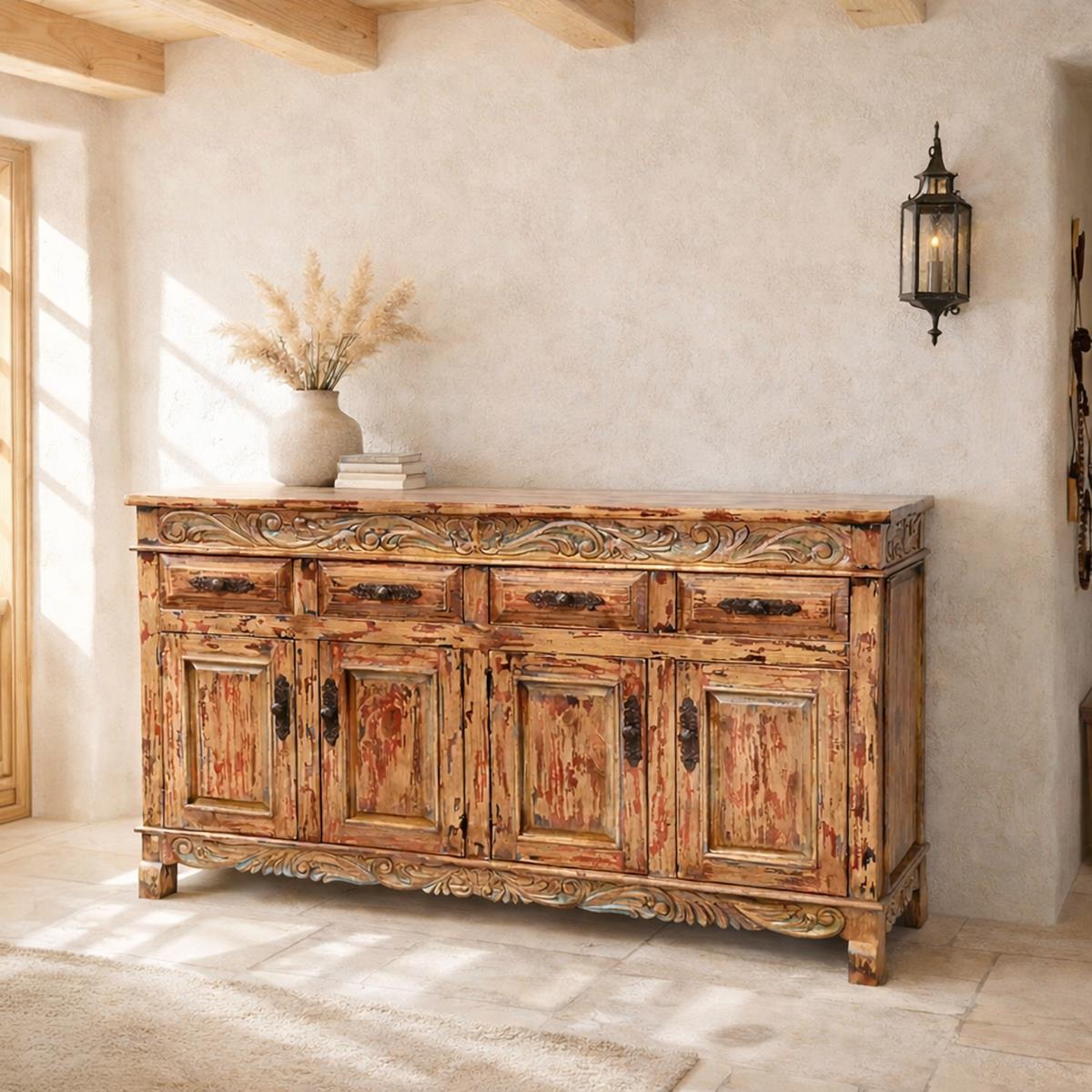 A rustic, distressed wooden sideboard cabinet with carved details, set in a warm, sunlit room with a textured plaster wall and a black wall sconce.