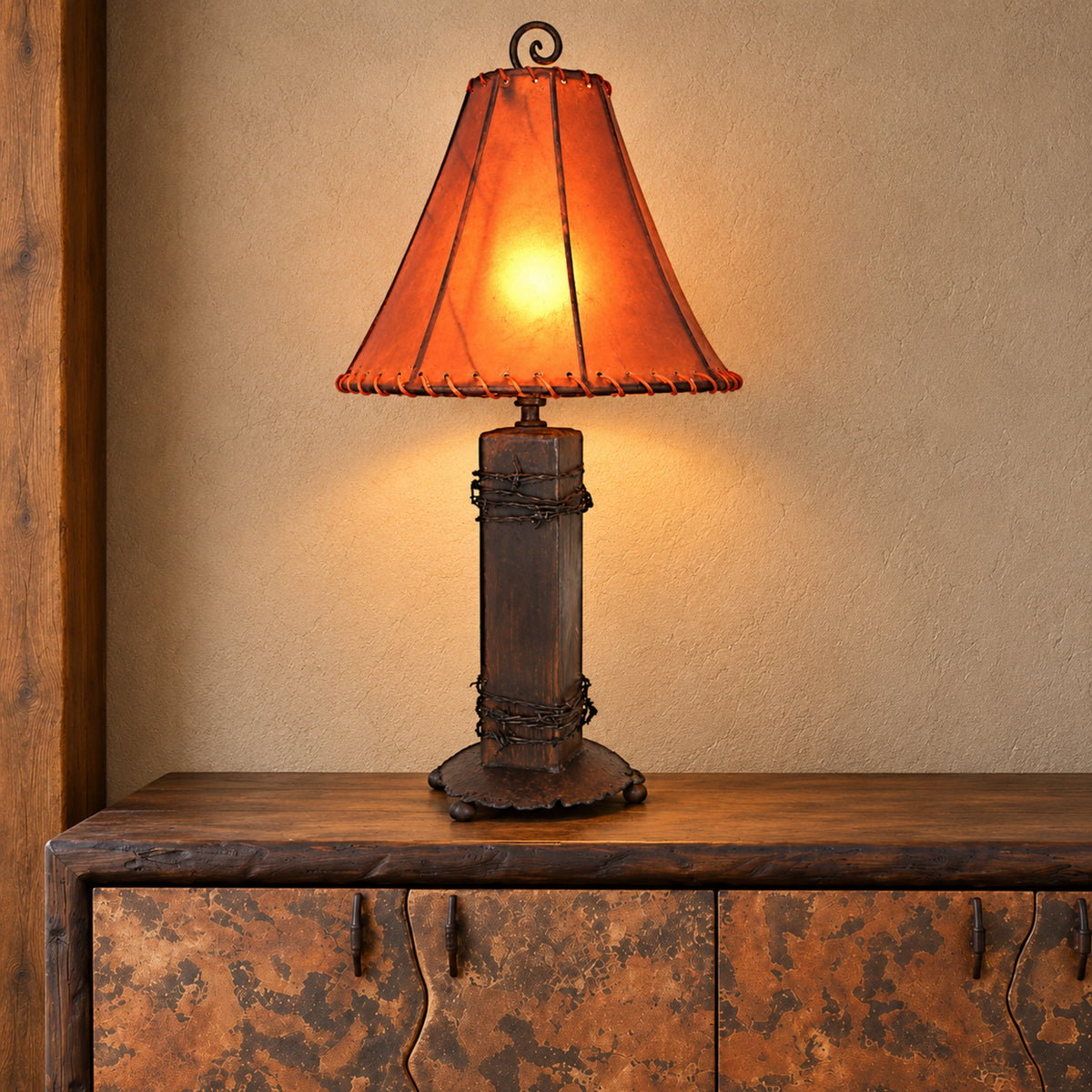 A rustic table lamp with a dark metal column base wrapped in barbed wire and a warm amber rawhide lamp shade, shown lit on a wooden and copper cabinet against a beige wall.