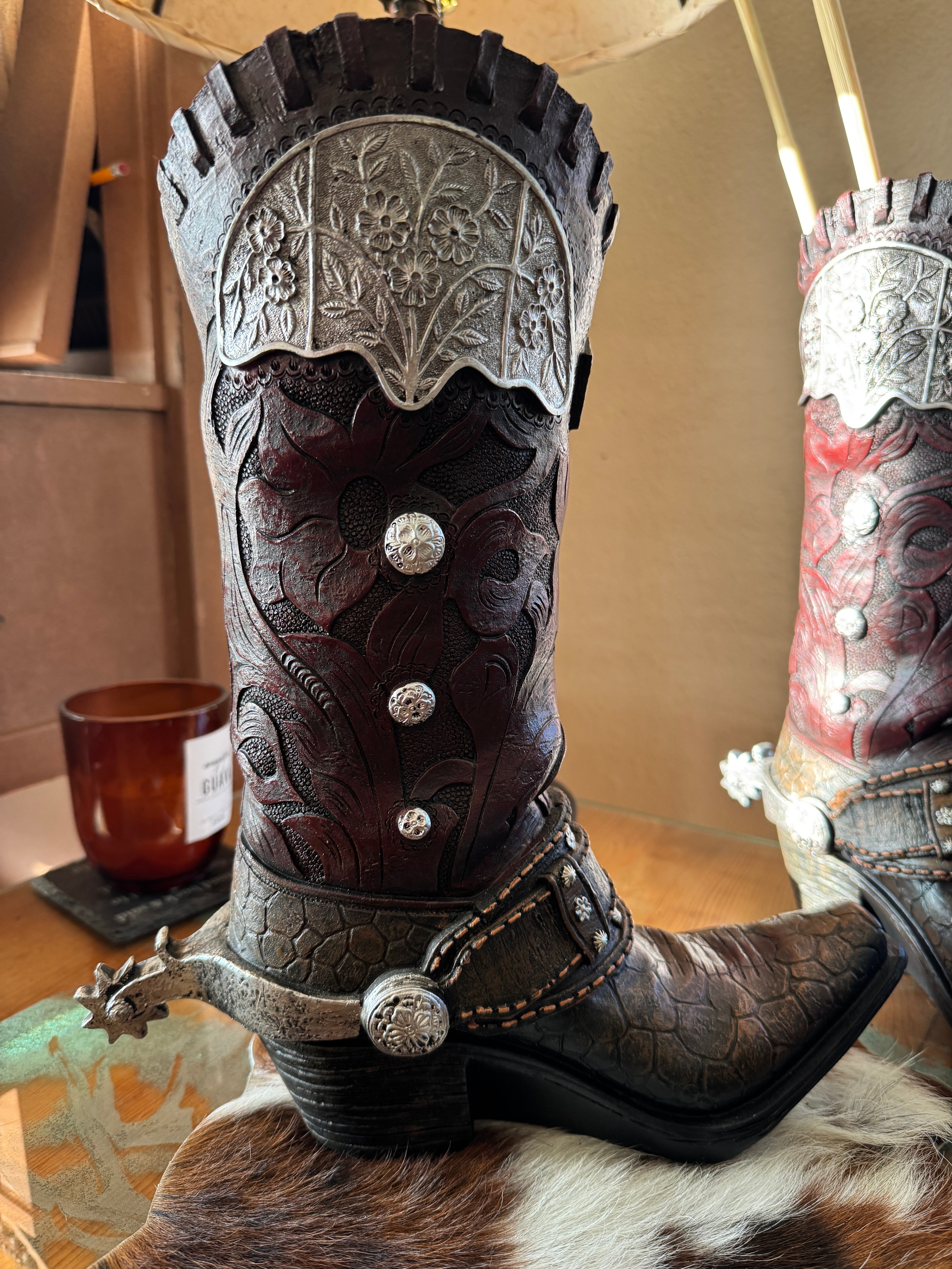 Close-up of a detailed cowboy boot vase with decorative elements on a cowhide mat over a wooden surface.