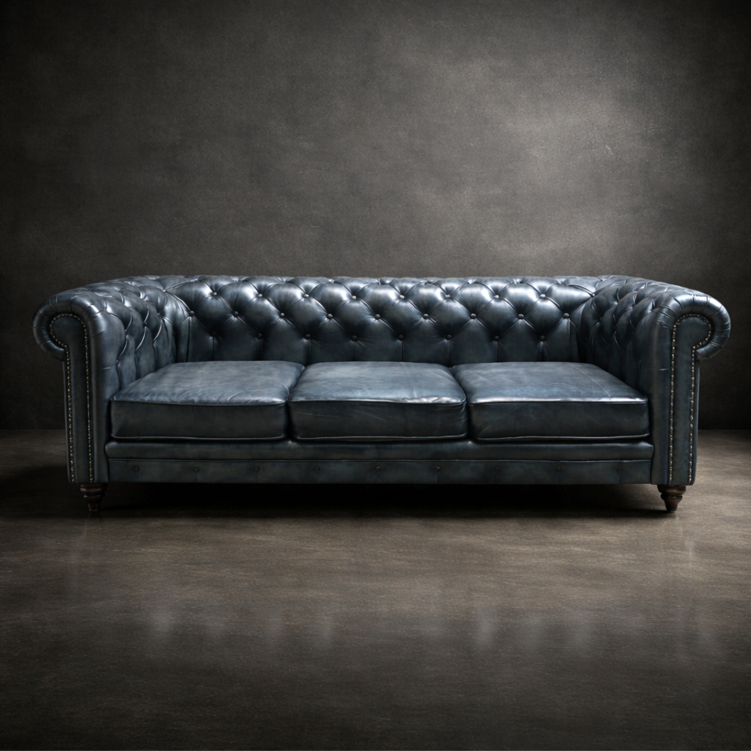 A dark blue tufted leather Chesterfield sofa with rolled arms and nailhead trim, photographed against a dark textured studio backdrop.