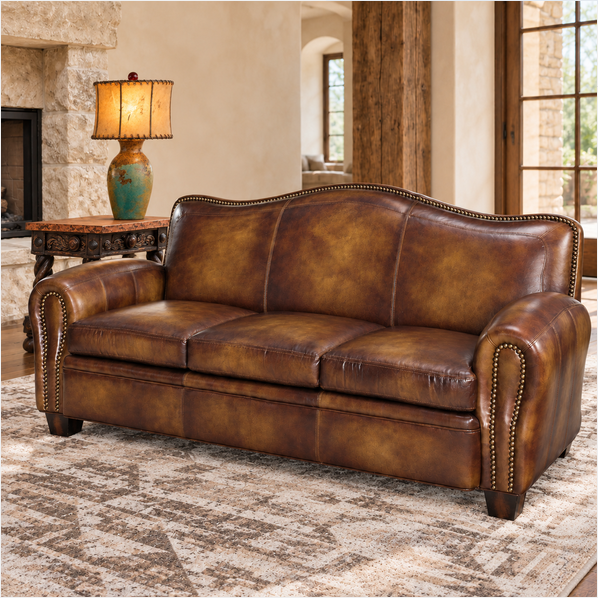 A brown burnished leather sofa with nailhead trim, shown in a rustic living room setting next to a side table with a lamp.