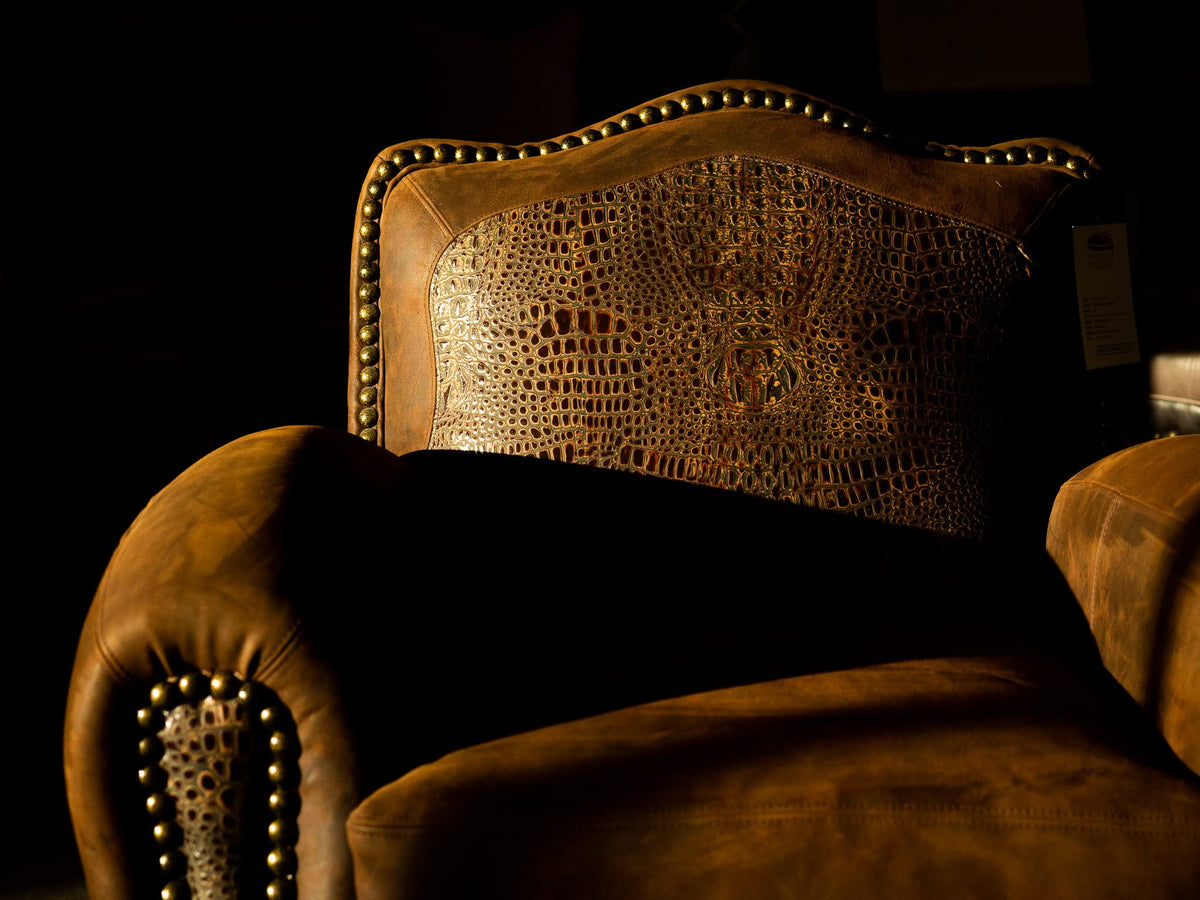 A close-up of a brown leather armchair with a crocodile-embossed backrest panel and brass nailhead trim, photographed in dramatic low light against a dark background.