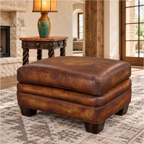 A square brown leather ottoman with a distressed finish and dark wooden legs, shown in a styled living room setting on a patterned rug next to a side table with a lamp.