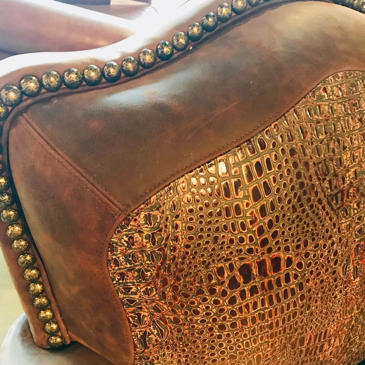 Close-up of a brown leather upholstered chair or sofa arm with a metallic gold embossed crocodile pattern panel and gold nailhead trim.