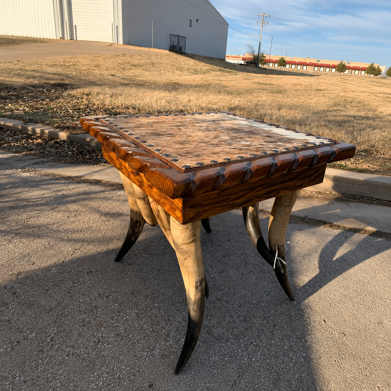 A rustic western square end table with a cowhide top, nailhead trim, carved wooden aprons, and four steer horn legs, photographed on a paved surface with a grassy lot and building in the background.