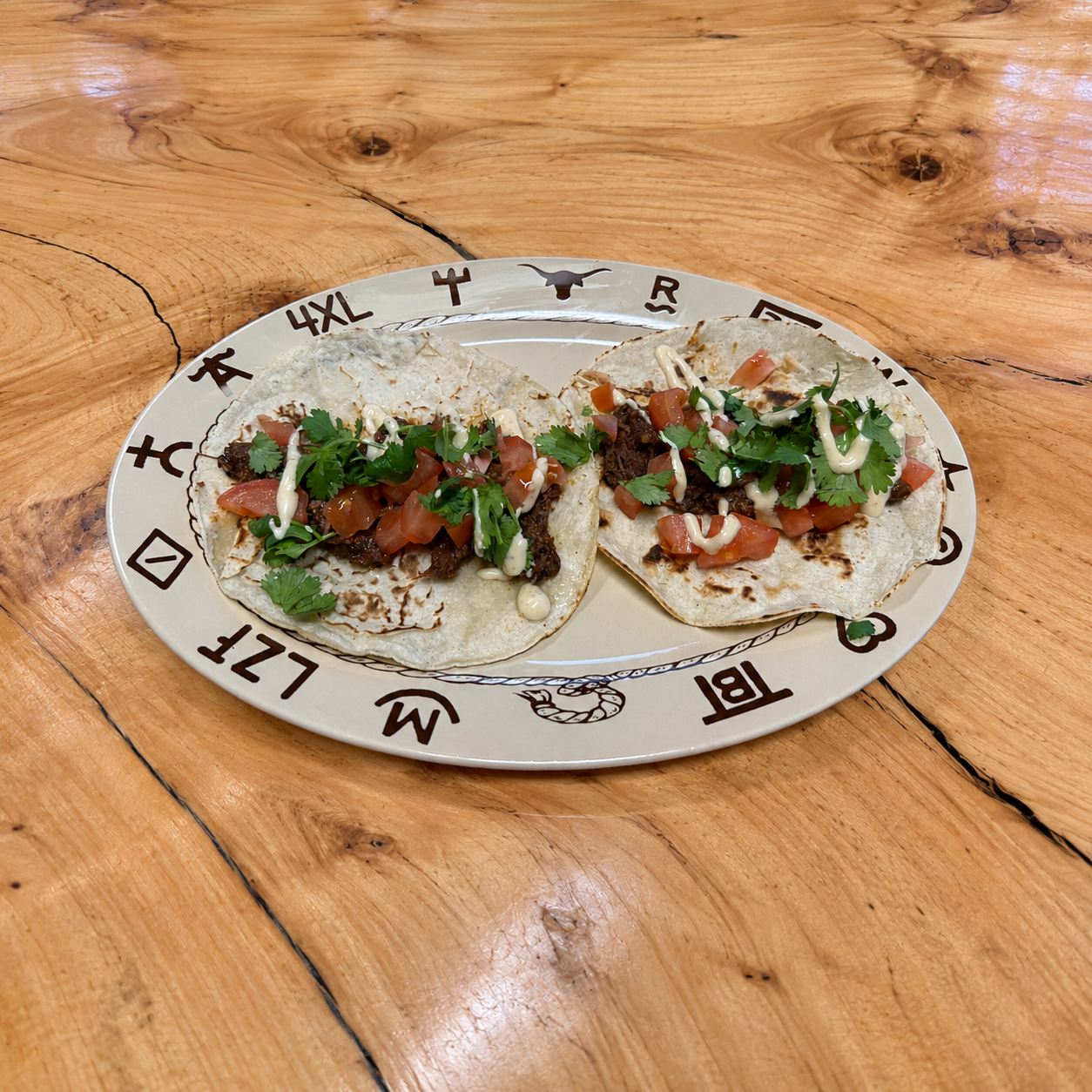 Two soft corn tacos topped with diced tomatoes, cilantro, and a white sauce, served on a beige plate with dark brown ranch brands and rope images, placed on a wooden table.