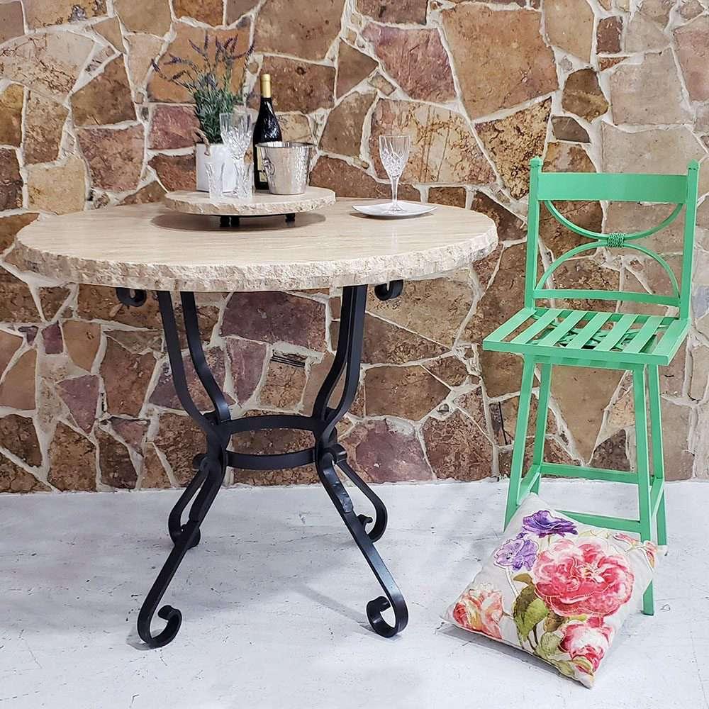 Round stone table with black metal legs against a stone wall, accompanied by a green chair and floral pillow - Your Western Decor