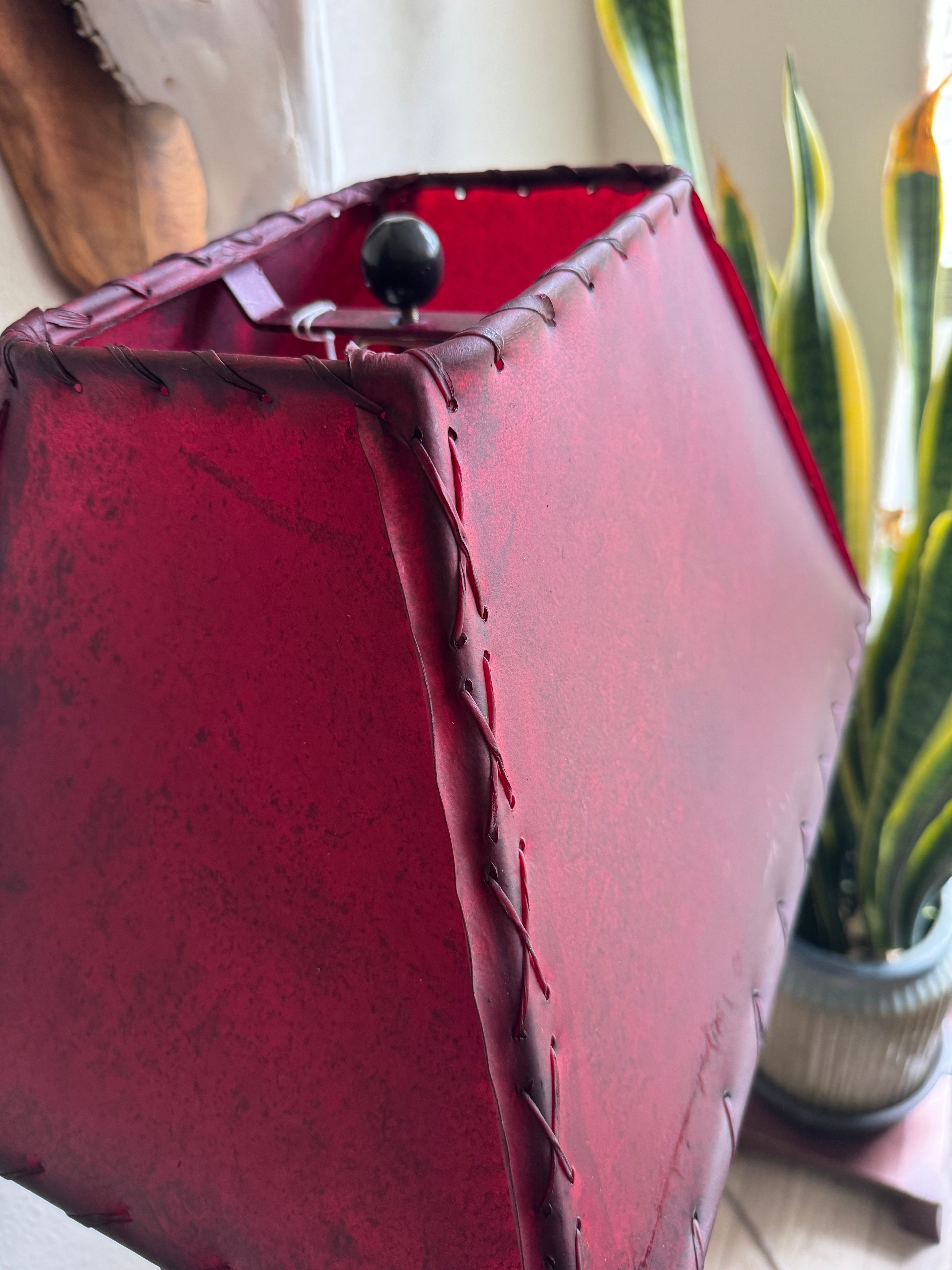 Close-up of a red leather lampshade with stitching details, blurred plant in the background