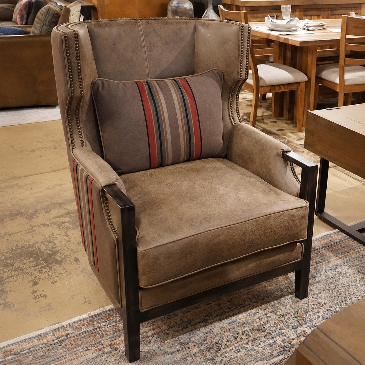 A brown wingback armchair with dark wood legs and nailhead trim, shown in a furniture showroom. The chair is fitted with a matching brown striped accent pillow featuring red, blue, and beige stripes.