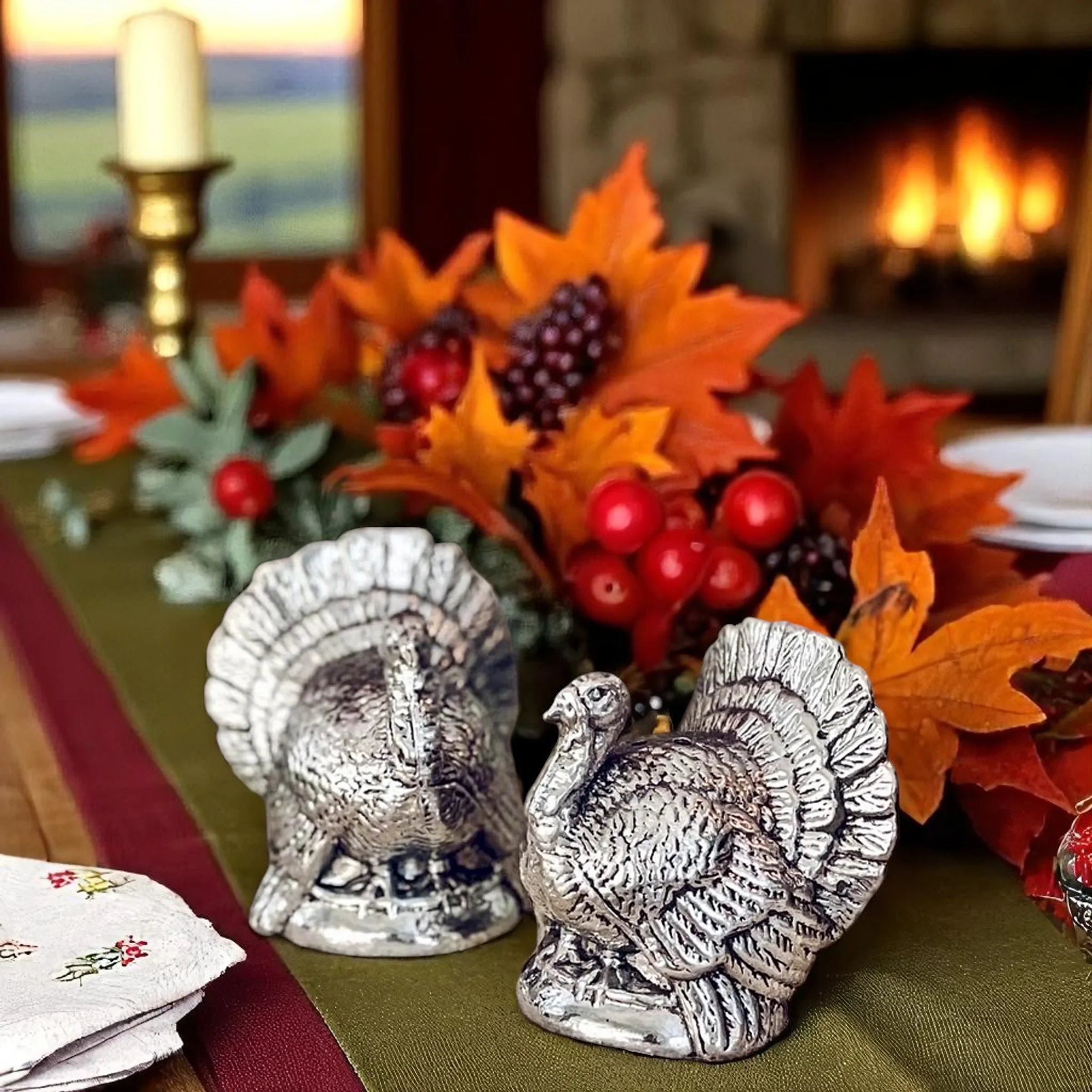 Two silver turkey figurines on a table with autumn leaves and berries, fireplace in the background.