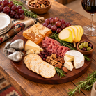 A rustic wooden charcuterie board with aluminum pear embellishment arranged on a wooden table with cheeses, crackers, cured meats, nuts, grapes, pear slices, olives, honeycomb, rosemary, and a glass of red wine nearby.