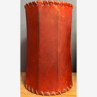 Red leather cylindrical lamp shade with whip stitching on a wooden surface and gray background