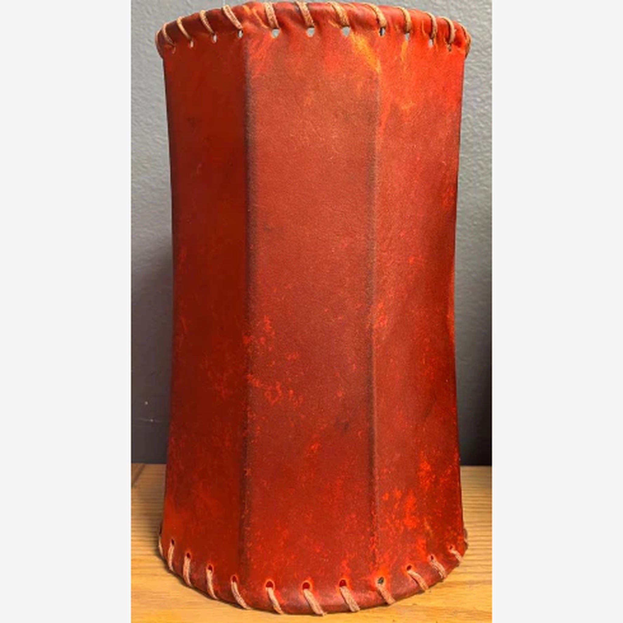 Red leather cylindrical lamp shade with whip stitching on a wooden surface and gray background