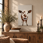 A vertical canvas wall art print depicting a brown and white cow, styled in a warm, rustic living room setting above a wooden console table with a vase, books, and candles.