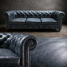 A dark blue tufted leather Chesterfield sofa shown in a studio setting, with a close-up of the matching leather upholstery in the foreground.