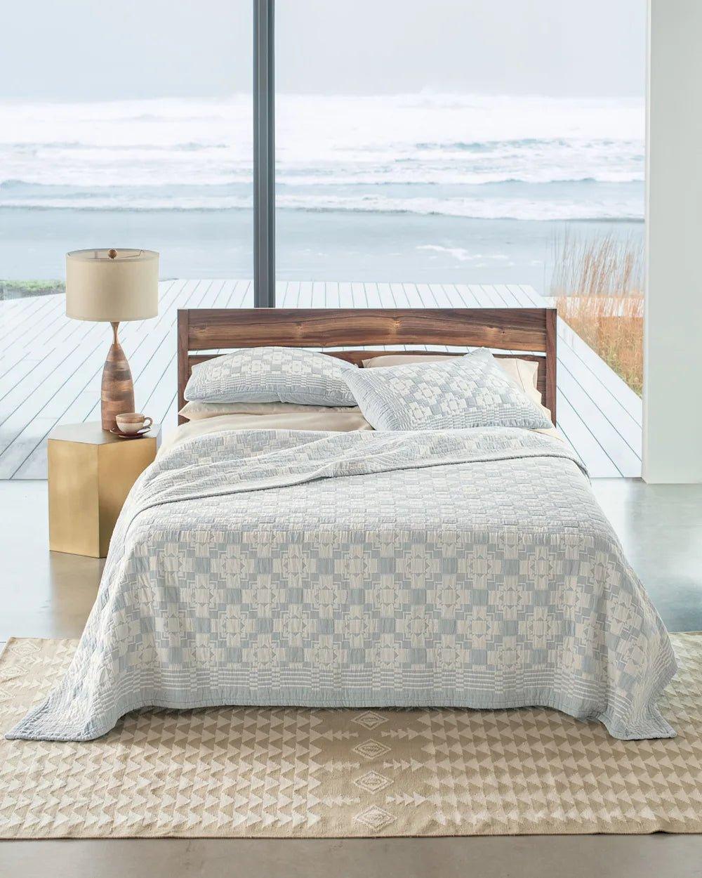 Bed with wooden headboard and light blue patterned quilt in a room with large windows showing a view of water.