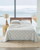 Bed with wooden headboard and light blue patterned quilt in a room with large windows showing a view of water.