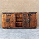 A rustic wooden sideboard cabinet with distressed brown wood, dark metal hardware, and an open center shelf, set against a textured beige wall and concrete floor.