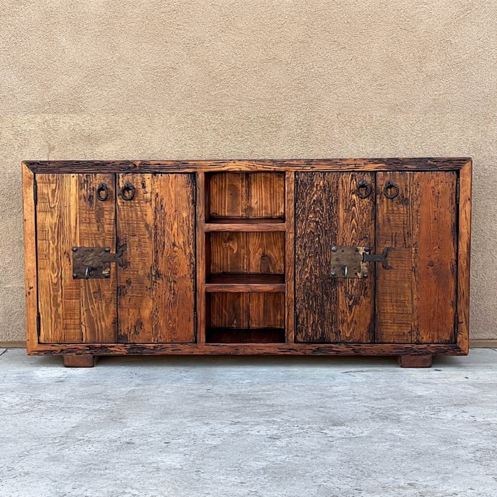 A rustic wooden sideboard cabinet with distressed brown wood, dark metal hardware, and an open center shelf, set against a textured beige wall and concrete floor.