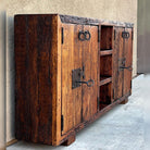 A rustic wooden sideboard cabinet with dark stained grain, two doors featuring antique metal ring pulls and latch hardware, and open shelving in the center, photographed against a stucco wall and concrete floor.