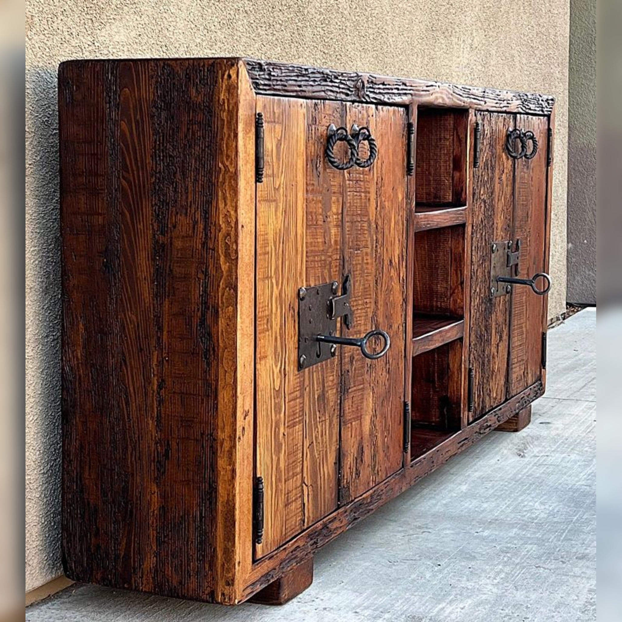 A rustic wooden sideboard cabinet with dark stained grain, two doors featuring antique metal ring pulls and latch hardware, and open shelving in the center, photographed against a stucco wall and concrete floor.