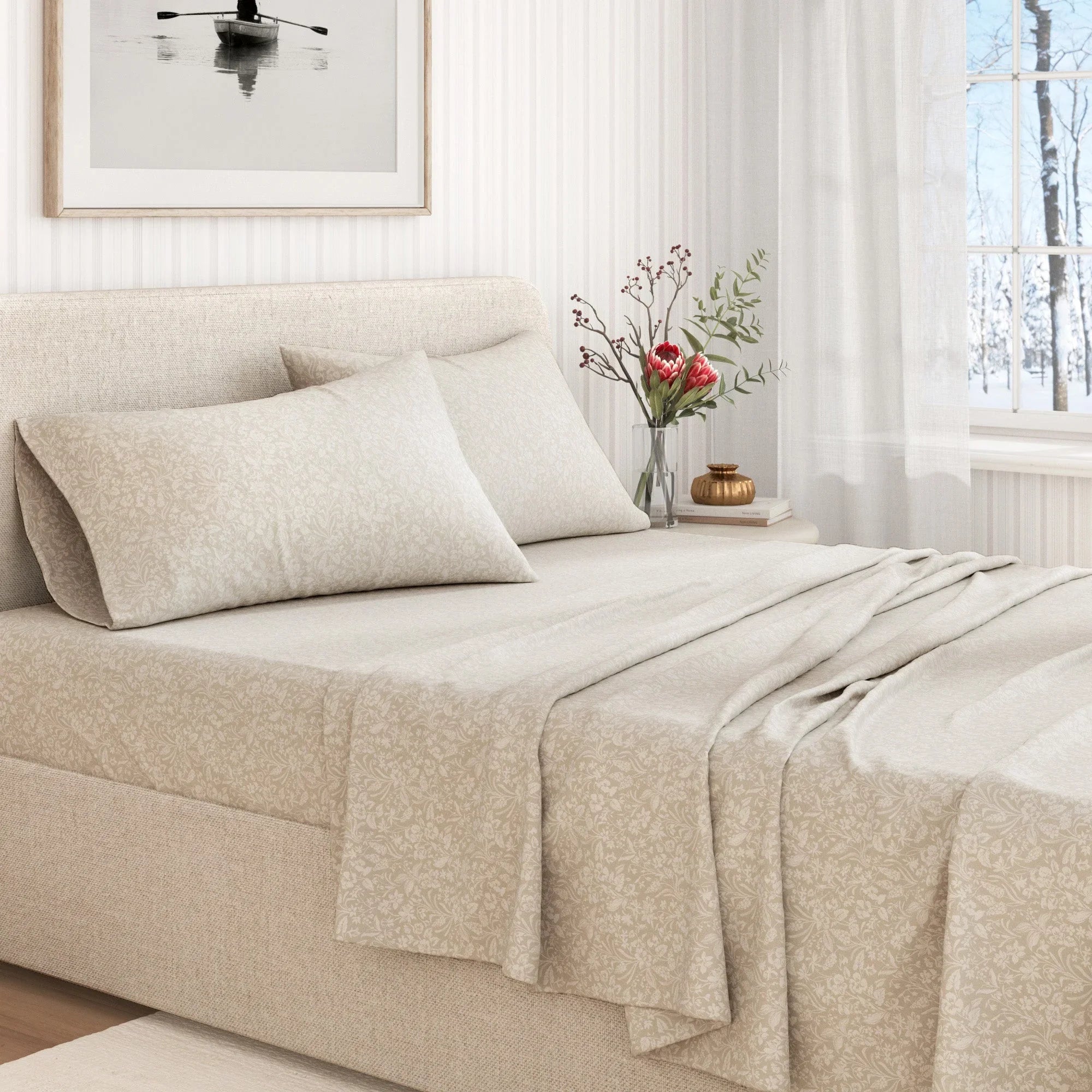 Beige bedspread and pillows on a bed in a bright bedroom with a vase of flowers on a nightstand.