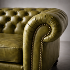 Close-up of an olive green tufted leather sofa arm with brass nailhead trim, photographed in a studio setting.