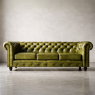 An olive green tufted leather Chesterfield sofa with nailhead trim and dark wooden legs, set against a neutral gray concrete wall and floor.