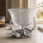 A clear glass bowl with a sculpted silver base featuring antlers, pinecones, and flowers, displayed on a tabletop in a softly lit interior setting.